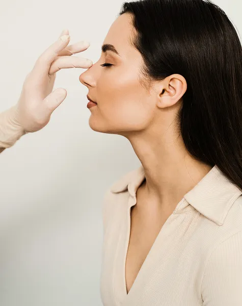 Mulher de perfil, enquanto uma mão com luva examina delicadamente o seu nariz.