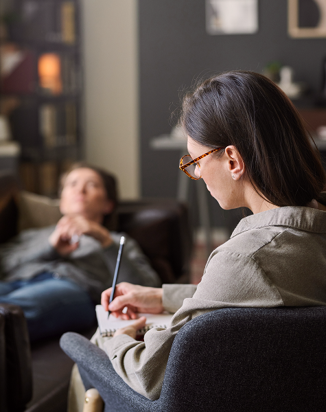 Psicóloga a tirar notas durante uma sessão de terapia, com o paciente deitado num divã ao fundo do consultório.